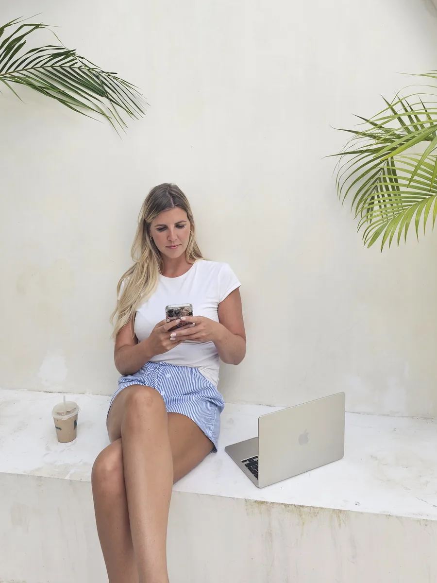 Lotti mit Handy und Laptop vor einer weißen Wand mit Palmen